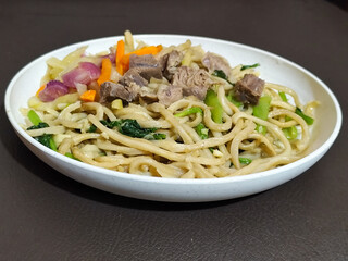 Rich, flavorful stir-fried udon noodles served on a white plate, accompanied by tender pieces of beef, sliced ​​fresh carrots and cabbage, and green mustard greens, create a delicious and appetizing d
