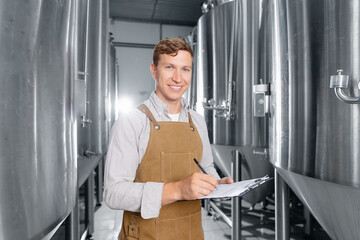 Young caucasian male brewer in brewery with fermentation tanks and clipboard, Quality control...