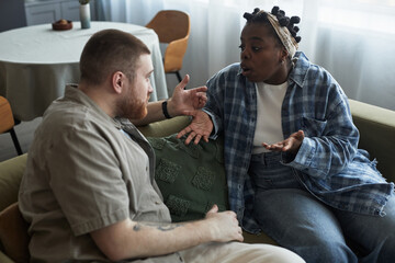 Young adult Caucasian man and young adult Black woman sitting on sofa having intense argument, both...