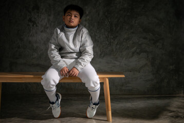 A boy in fencing gear sitting on a bench, ready for the épée competition