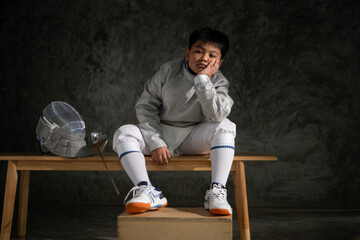 A boy in fencing gear sitting on a bench, ready for the épée competition