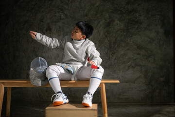 Young boy in fencing gear sitting on a bench, examining his épée sword with focus