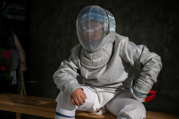 A boy in fencing gear sitting on a bench, ready for the épée competition