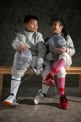 Two child in fencing gear sit on a bench, smiling at each other