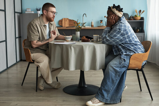 Caucasian young adult man and Black young adult woman sitting at round table facing each other, gesturing and listening with serious expressions, engaging in difficult conversation