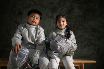 Two child in fencing gear sit on a bench, smiling at each other