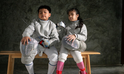 Two child in fencing gear sit on a bench, smiling at each other