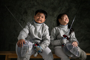 Two child in fencing gear sit on a bench, smiling at each other