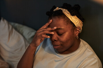 Black woman young adult sitting on bed touching forehead with closed eyes showing stress or...