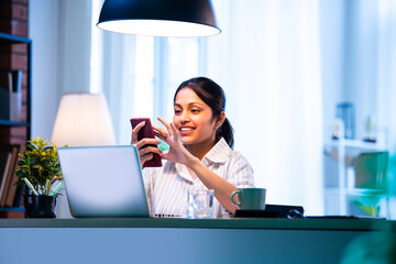 Indian female employee checks mobile at cozy desk, multitasking and smiling during office hours