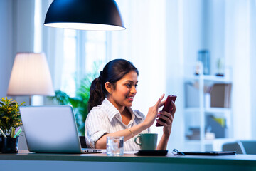 Indian female employee checks mobile at cozy desk, multitasking and smiling during office hours