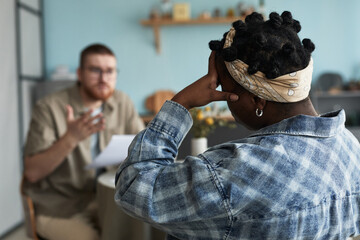 Black woman with headscarf sitting with back to camera, holding head while Caucasian young adult...