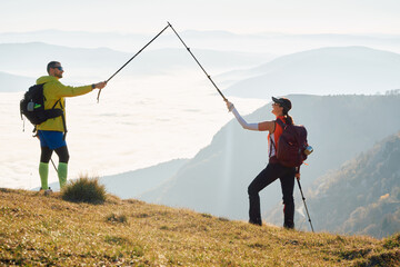 Couple hikers trek up a rocky mountain trail, enjoying a scenic view above the clouds, with majestic peaks in the background.