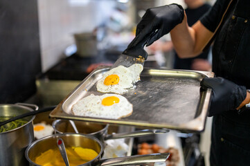 Chef plating fried eggs on a metal tray in a busy professional kitchen, surrounded by pots, pans,...