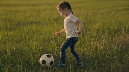 Little child runs across the green football field playing football at sunset in the sky, kid kicks...