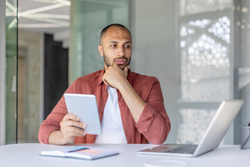 A thoughtful man at a desk holds a tablet, pondering ideas while working on a laptop in a modern...