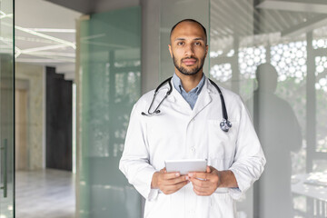 A doctor, wearing a stethoscope and lab coat, stands in an office holding a tablet. He appears calm...