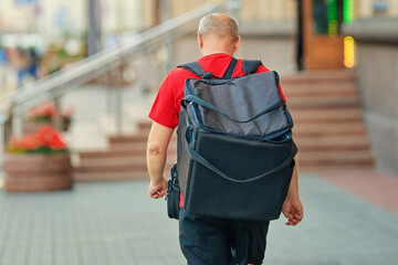 Aged deliveryman with food bag on his back strides down city street. Elderly food delivery worker wearing backpack moves through city streets during work