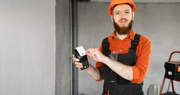 Happy builder or engineer in hardhat holding pos terminal and credit card for contactless payment standing at construction site - Powered by Adobe