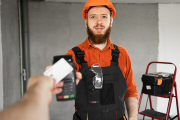 Bearded builder or repairman wearing hardhat hold wireless bank payment terminal client pay for...