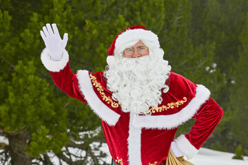 Santa Claus with a sack of gifts stands in a snowy forest and waves his hand in greeting.