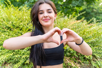 young beautiful woman showing heart sign outdoors in front of green bush