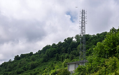 Telecommunication tower amidst lush hill. A telecommunication tower rising above a dense green hillside under a cloudy sky. Use for themes related to connectivity and infrastructure.