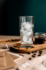 Glass filled with ice cubes stands next to a small measuring cup of espresso. Preparing iced latte in a cozy setting with scattered coffee beans.