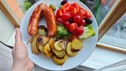 Homemade dinner plate with grilled sausages, roasted potatoes, and fresh salad with cherry tomatoes and olives. Represents hearty meal, comfort food, and colorful home cooking concept