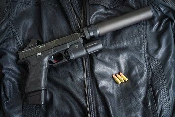 A tactical pistol with a silencer, red dot sight, and flashlight lies on a black leather jacket...