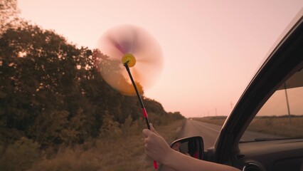 free girl waving a pinwheel out of the car window in wind, travel adventure road at pink sunset,...