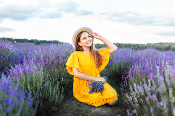 Beautiful young girl in straw boater hat and a yellow dress collects lavender on lavender field....