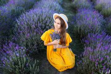 Beautiful young girl in straw boater hat and a yellow dress collects lavender on lavender field....