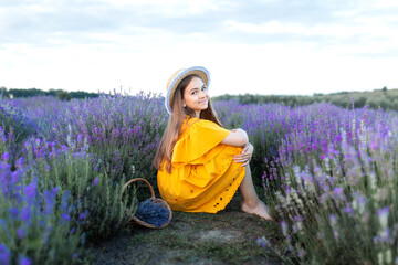 Beautiful young girl in straw boater hat and a yellow dress collects lavender on lavender field....