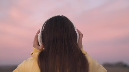 put on headphones on your head, a beautiful pink sunset in sky, listen to music tracks in speakers with your ears, get musical pleasure and relax, record audio at sunset, diversify your leisure time.