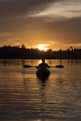 A man paddles a kayak on a tropical island with a sunset in the background. A man enjoys the sunset while playing kayak
