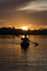 A man paddles a kayak on a tropical island with a sunset in the background. A man enjoys the sunset while playing kayak