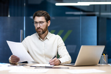 A serious young male businessman is sitting in an office at a desk, working on a laptop and with...