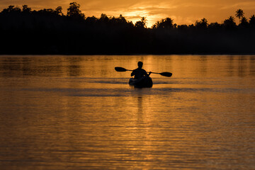 A man paddles a kayak on a tropical island with a sunset in the background. A man enjoys the sunset while playing kayak