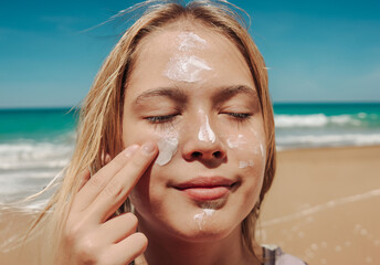 Teen applying sunscreen on face, arm, and leg under direct sun. Summer skincare, blue sky, beach background, and beige hammock. Sun protection routine, outdoor wellness