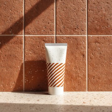 A white tube with orange stripes and speckled accents sits on a light counter against a terracotta tile wall, bathed in sunlight