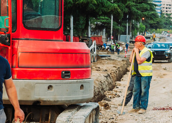 Construction workers repairing city infrastructure with heavy equipment and red polypropylene pipes. Urban utility maintenance, excavation, and roadwork in progress
