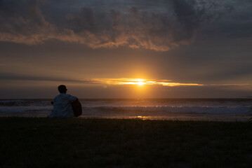 A young man sits and plays guitar on a tropical beach at sunset. A happy man enjoys the sunset while playing guitar on the beach