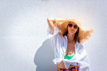 Confident woman wearing sun hat and sunnies agains standing at white wall in the sunshine