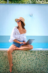 A woman sitting on the edge of the pool using a laptop