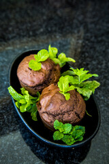 Two chocolate mint flavoured muffins, decorated with mint leaves, in a small, black elongated plate on a shiny stone table top.