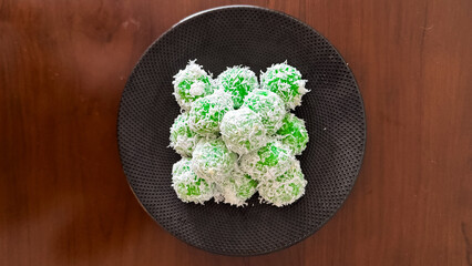 Traditional Indonesian klepon, green sticky rice balls with palm sugar, coated in shredded coconut, served on a black plate over a wooden table.