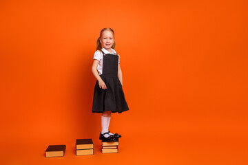 Young schoolgirl in uniform standing on stacked books with orange background, embracing childhood...