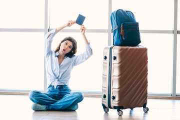 Charming woman sitting on the floor at an airport, holding a passport, waiting with her suitcase...