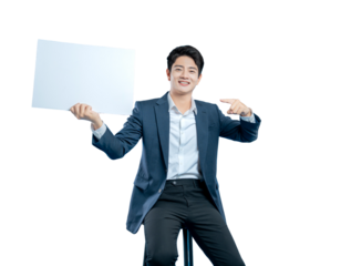 A smiling young man in a suit sits on a stool, pointing to a blank white placard he is holding, perfect for showcasing a message.
